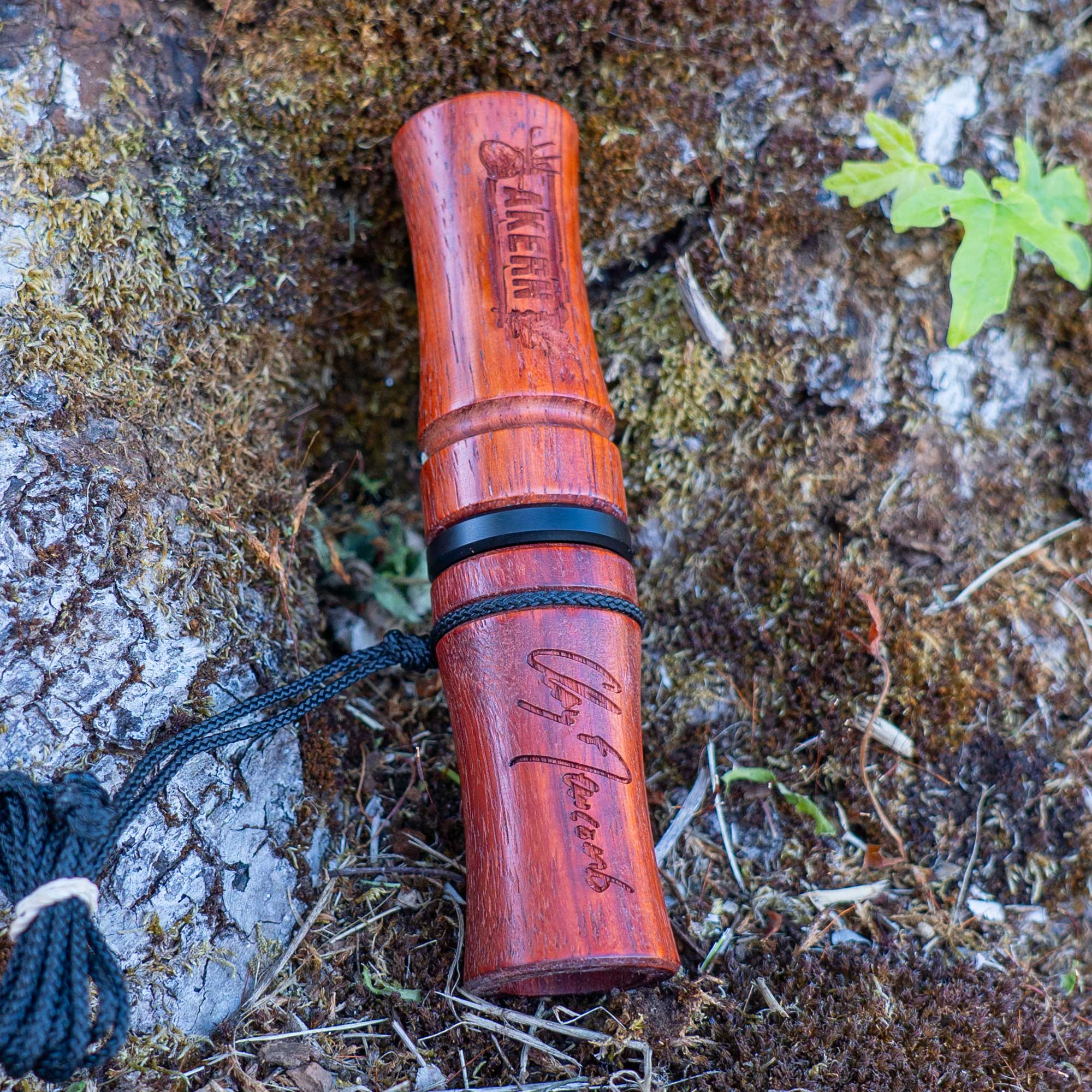 A padauk wood deer call with "Akern" text and Clay Newcomb signature on a mossy stump in the woods